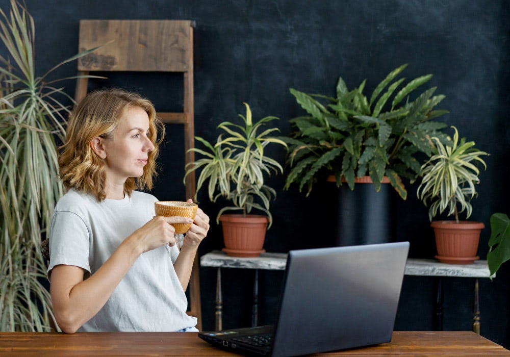 A person holds a mug while sitting in front of a laptop on a wooden desk, surrounded by various potted plants against a dark wall with a wooden ladder.